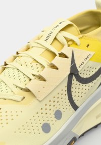 Chaussure de trail Nike de couleur jaune pâle, avec un dessus en mesh, des accents texturés, des perforations et un logo gris. Les lacets sont de couleur beige clair.