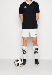 Svart kortärmad v-ringad tröja, vit krage, kombinerad med vita shorts och svarta fotbollssockor med ett svart mönster. Svart fotboll vid fötterna.