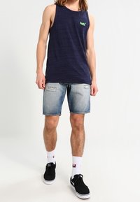 Navy blue sleeveless tank top with green logo, paired with light blue denim shorts featuring frayed hems. Black sneakers and white socks.