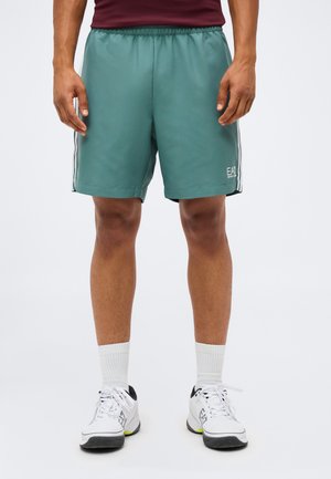Mand iført turkis sportsshorts med sidelommer, hvide sportsstrømper og hvide sneakers med sorte detaljer.