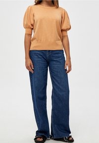 Maglione a maniche a sbuffo di un arancione chiaro con orlo a coste, abbinato a jeans a gamba larga blu scuro e sandali neri. Design semplice e texture liscia.