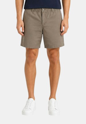 Uomo con pantaloncini kaki con vita elastica e bottone, abbinati a sneakers bianche e una maglia scura, in piedi su uno sfondo semplice.