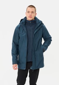 Giubbino impermeabile blu scuro con cappuccio, dotato di tasche con zip e finitura liscia, indossato sopra un maglione navy con texture.