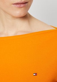 Oranges Baumwoll-T-Shirt mit weitem Ausschnitt. Verfügt über ein kleines rot, weiß und blaues Logo auf der Brust. Glatte Textur und figurbetontes Design.