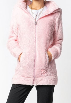 Femme portant une veste rose clair duveteuse à fermeture éclair avec capuche, mains dans les poches, chemise blanche en dessous, et colliers en or superposés.