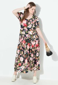 Frau in einem schwarzen, floral gemusterten, knopfgeschlossenen Midikleid, die eine zweifarbige, geflochtene Handtasche hält, weiße Plateauschuhe trägt und vor einer weißen Wand posiert.