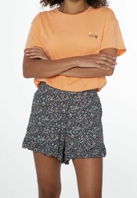 Orange bomullströja med en liten logotyp på bröstet, parat med svarta shorts med blommönster i flera färger och scalloped nederkant.