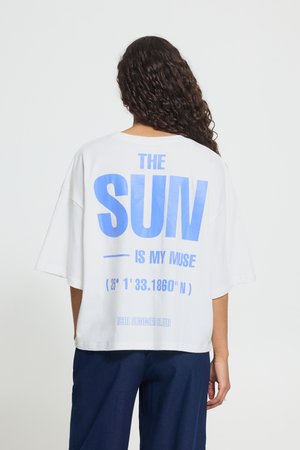 Isik pikkade lokkis juustega kannab valget üle suureks kasvanud t-särki, millel on suur sinine tekst "THE SUN IS MY MUSE", ja tumesiniseid pükse, näoga eemal ühevärvilise tausta ees.