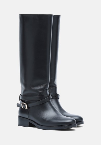 Paire de bottes noires en cuir montant jusqu'aux genoux avec des talons bas et des brides décoratives à la cheville avec boucles argentées.