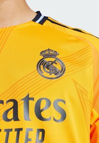 Maglietta da calcio gialla con rifiniture nere, caratterizzata dal logo del Real Madrid in argento e dal testo in grassetto "Emirates". Design tessurizzato con strisce diagonali.