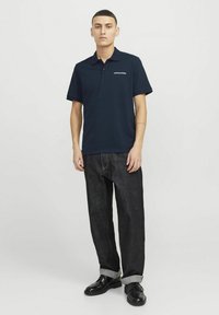 Mørkeblå polo med krave, med et hvidt logo. Båret med mørke jeans og sorte sko, stående foran en neutral baggrund.