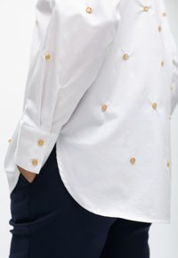 Camicia bianca con bottoni decorativi dorati, maniche lunghe e orlo curvo, abbinata a pantaloni blu scuro. Tessuto liscio con una finitura rigida.