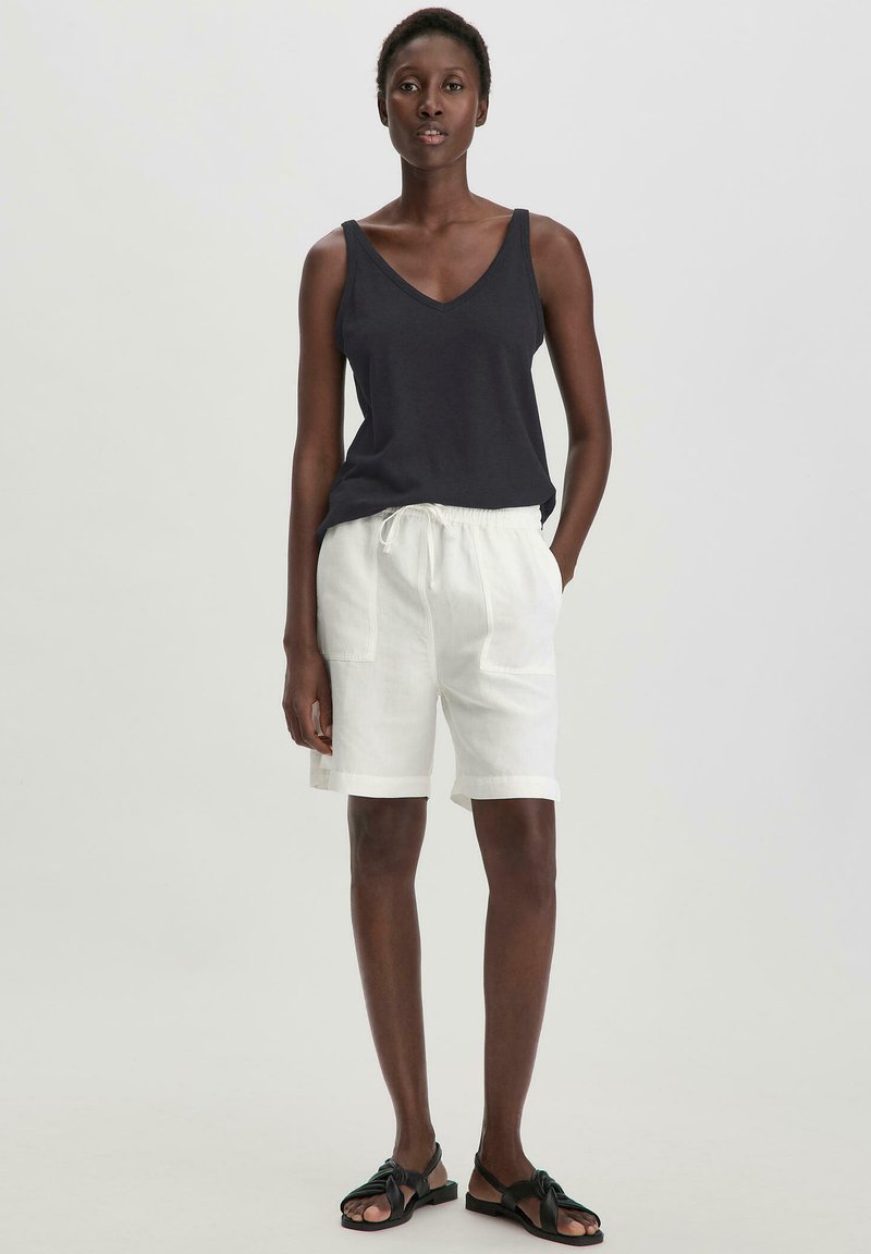 Dunkelblaues ärmelloses Oberteil kombiniert mit weißen Leinen-Shorts mit einem Kordelzugbund und Seitentaschen. Getragen mit schwarzen Riemchensandalen.