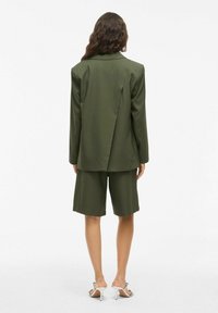 Blazer cintré vert olive avec un design à dos fendu, associé à un short assorti. Tissu texturé, manches de longueur moyenne et poches latérales.