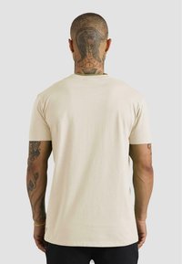 Muž s tetováním hada na holé hlavě a krku, oblečený v béžovém tričku a černých kalhotách, zobrazený zezadu na neutrálním pozadí.