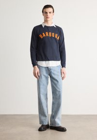 Sweater azul-marinho com "BARBOUR" em letras laranja, sobre uma camisa branca com colarinho. O modelo usa jeans azul-claro e botas pretas.