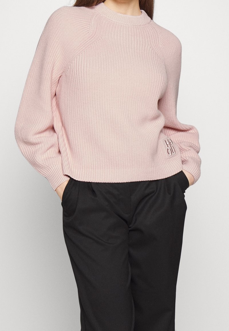 Femme portant un pull en maille côtelée rose clair avec des manches bouffantes et un pantalon noir, mains dans les poches, de la nuque aux hanches.