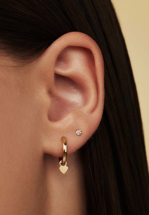 Boucle d'oreille en or avec un charm en forme de cœur, associée à une petite clou en argent ornée d'une pierre précieuse transparente. Gros plan sur l'oreille et les boucles d'oreilles.