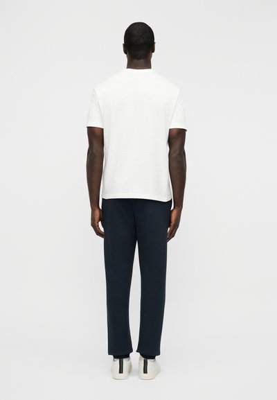 Λευκό βαμβακερό μπλουζάκι με κοντά μανίκια, όψη από πίσω· συνοδεύεται από ναυτικό μπλε παντελόνι και λευκά αθλητικά παπούτσια με πράσινες και μαύρες ρίγες στη φτέρνα.