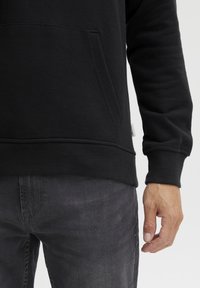 Felpa nera con tasca frontale, polsini e orlo a costine, abbinata a jeans grigi scuri. Il materiale sembra morbido e dalla consistenza interessante.