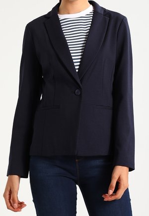 Frau trägt einen taillierten marineblauen Blazer über einem schwarz-weiß gestreiften Hemd und dunkelblaue Jeans, steht mit entspannt an den Seiten hängenden Händen.