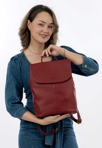 Sac à dos en cuir rouge avec une texture lisse, un rabat arrondi et une poignée supérieure. Bretelles ajustables et un subtil logo sur le devant.