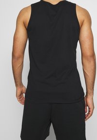 Un homme portant un débardeur noir et un short noir, vu de dos, se tenant contre un fond clair.