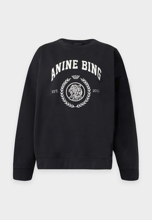 Osoba uśmiecha się, ma na sobie czarną bluzę „"ANINE BING"”, niebieskie szerokie dżinsy, czarne szpiczaste buty i trzyma małą czarną torebkę.