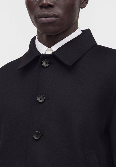 Close-up of a person wearing a black buttoned coat with a white collared shirt underneath against a plain background.