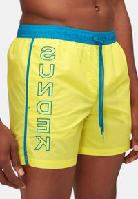 Bañador amarillo brillante con una cinturilla y cordón azul. Presenta un logo azul vertical a lo largo del costado. Textura ligera y suave.