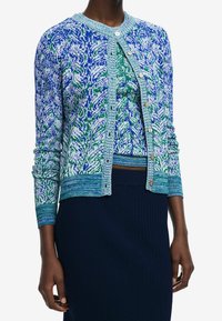 Lähikuva mallista, joka pukee ylleen sinivihreäkuvioisen neuletakin helmipainikkeilla, samansävyisen topin sekä laivastonsiniset resorihameen.