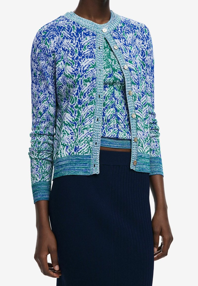 Lähikuva mallista, joka pukee ylleen sinivihreäkuvioisen neuletakin helmipainikkeilla, samansävyisen topin sekä laivastonsiniset resorihameen.