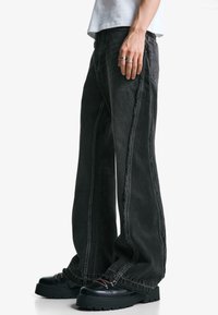 Široke črne jeans hlače s kontrastnim šivanjem, robustno teksturo in strganimi robovi, v kombinaciji z masivnimi črnimi čevlji.