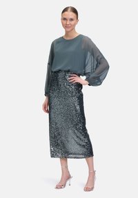 Top lungo in chiffon a maniche lunghe color teal abbinato a una gonna midi in paillettes argento. La modella indossa tacchi a spillo argentati. Estetica pulita e minimalista.