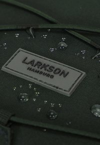Sötétzöld szövet, amelynek texturált felülete van, szürke gumibélyegzővel, amelyen a "LARKSON HAMBURG" felirat látható. Vízcseppek a anyagon.