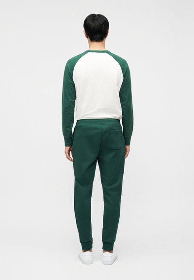 Άτομο που στέκεται γυρισμένο, φοράει λευκό και πράσινο μπλουζάκι με μακριά μανίκια, σκούρα πράσινα παντελόνια jogging και λευκά αθλητικά παπούτσια σε απλό φόντο.