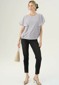 Donna con blusa a maniche svolazzanti lilla, jeans skinny neri, tacchi beige, che tiene una borsa beige, mentre guarda di lato su uno sfondo neutro.