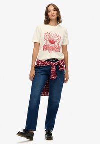 Beżowy t-shirt z grafiką w czerwonym kolorze przedstawiającą góry, w połączeniu z ciemnoniebieskimi prostymi jeansami oraz czerwoną koszulą w kratę zawiązaną wokół talii. Czarne buty.