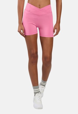 Mujer con pantalones cortos deportivos rosas de tiro alto, calcetines grises a rayas y zapatillas blancas, de pie con las piernas ligeramente cruzadas.