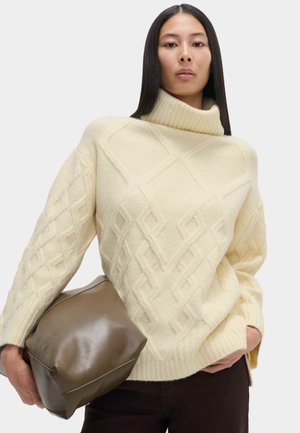 Cremfarbener Pullover mit hohem, geripptem Kragen und Rautenmuster. Das Modell hält eine braune, weiche Ledertasche. Glatte Textur, lässige Passform.