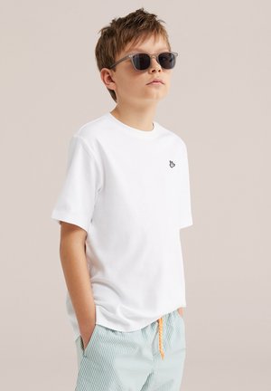 Junge mit dunkler Sonnenbrille, weißem T-Shirt mit kleinem Hand-Logo und hellblauen gestreiften Shorts mit orangem Kordelzug, stehend mit Händen in den Taschen.