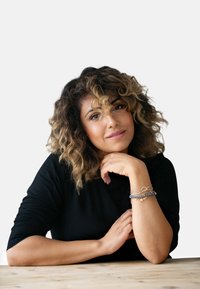 Femme aux cheveux frisés portant un haut noir côtelé, reposant son menton sur sa main. Elle a plusieurs bracelets de matériaux variés au poignet.