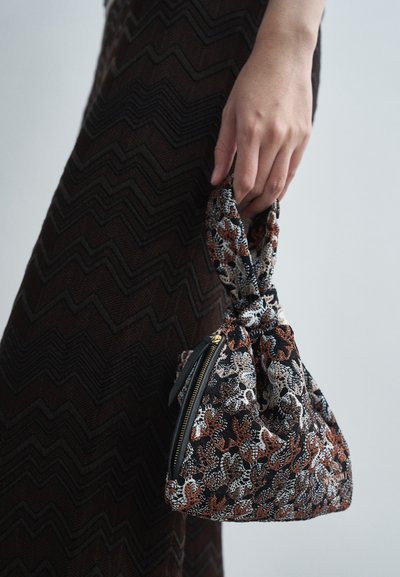 Χέρι που κρατάει μια μικρή υφασμάτινη τσάντα χειρός με λουλουδάτη κεντητική σε μαύρο, καφέ και μπεζ, πάνω από μια βραδινή φούστα με καφέ ζιγκ-ζαγκ σχέδιο.