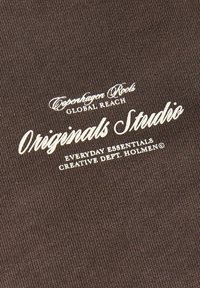 Tissu brun avec texte blanc indiquant Copenhagen Roots Global Reach Originals Studio Everyday Essentials Creative Dept. Holmen.