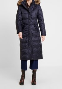 Tmavě modrý péřový kabát s kapucí lemovanou umělou kožešinou, předním zapínáním na zip a bočními kapsami. Má prošívanou strukturu a dlouhý střih.