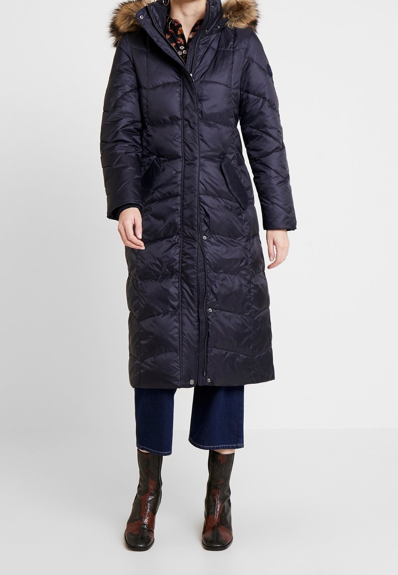 Tmavě modrý péřový kabát s kapucí lemovanou umělou kožešinou, předním zapínáním na zip a bočními kapsami. Má prošívanou strukturu a dlouhý střih.