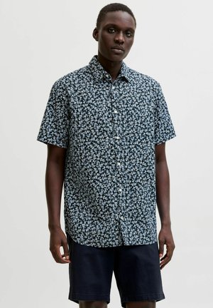 Fiatal férfi fekete-fehér virágos rövid ujjú gombos ingben és sötét rövidnadrágban, világos egyszínű háttér előtt áll.