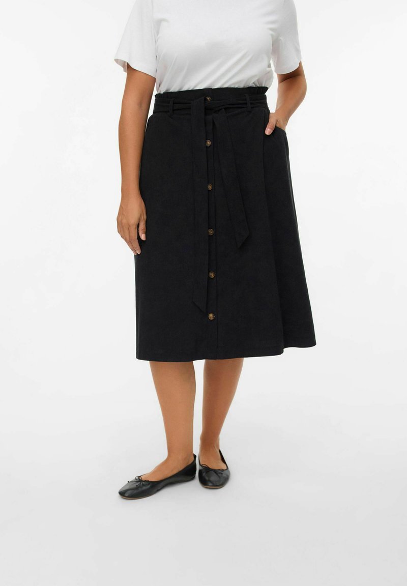 Jupe midi noire en tissu texturé, dotée d'une taille nouée, d'une fermeture à boutons sur le devant et de poches latérales. Portée avec un T-shirt blanc et des ballerines.