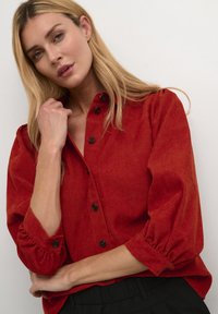 Chemise boutonnée en velours côtelé rouge avec des manches ballon et des boutons noirs, portée sur un pantalon sombre. Le tissu présente une finition texturée.