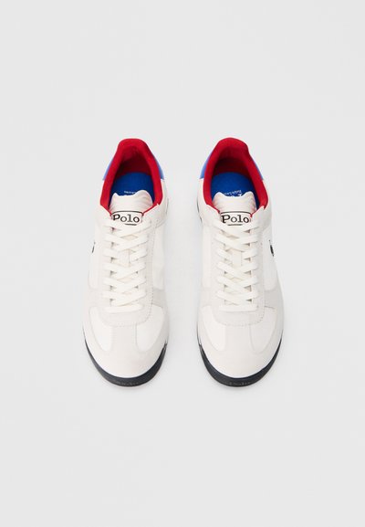Ténis brancos com parte superior em tecido texturizado, detalhes azuis e vermelhos no colarinho e sola de borracha preta. Logótipo "Polo" na língua.